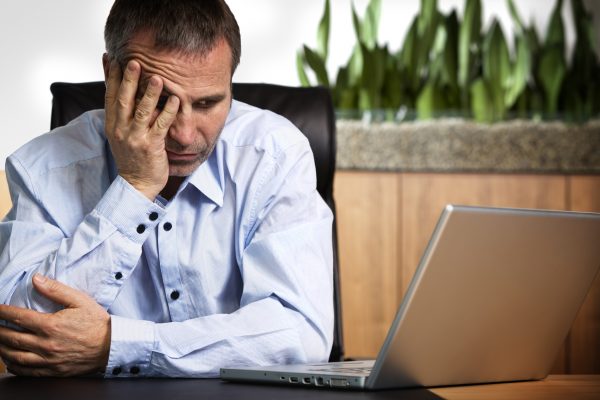 Senior business person in blue shirt sitting at office desk looking at laptop and being frustrated about computer or software crash.